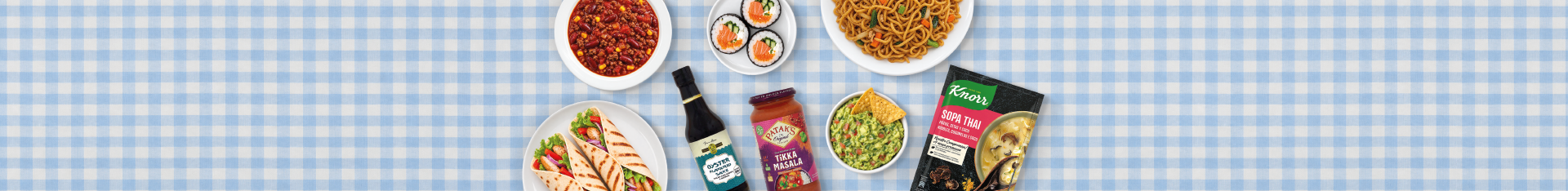 Imagen de cocina internacional con una selección de platos y productos de diferentes gastronomías del mundo. Incluye sushi, noodles orientales, chili con carne, wraps, guacamole, salsa tikka masala, salsa de ostras y sopa thai sobre un mantel de cuadros azules y blancos.